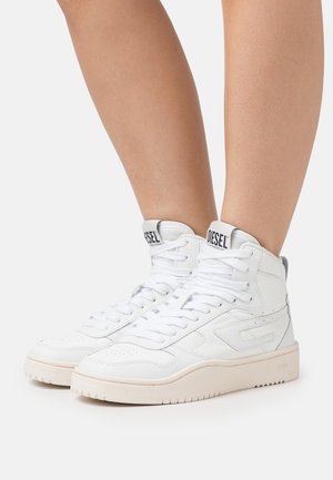 Hvide hvide høje sneakers fra Diesel med snørebånd, båret på bare ben mod en ensfarvet, lys baggrund.