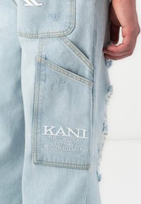 Light blue denim cargo pants with distressed details, featuring multiple pockets and white embroidered branding on the thigh.