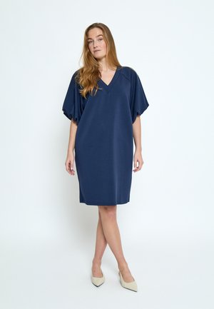 Mujer de cabello largo, vestida con un vestido azul marino holgado hasta la rodilla y tacones beige, de pie frente a un fondo blanco liso.