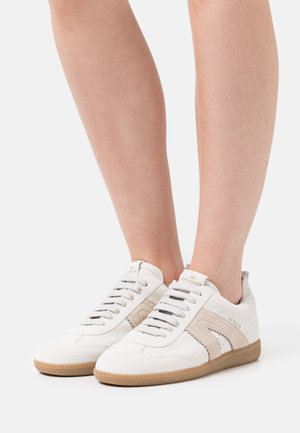 White suede sneakers with beige accents, featuring a lace-up design, textured sole, and subtle embossed branding at the heel.
