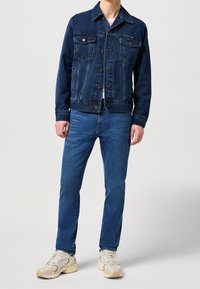 Homme portant une veste en jean bleu foncé, un jean bleu, une chemise blanche et des baskets blanc cassé, debout devant un fond uni.