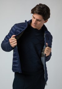 Giacca imbottita blu navy con texture trapuntata e fodera interna grigia, con chiusura a zip e colletto alto, indossata sopra un maglione scuro.