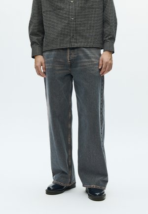 Džíny s rozšířenými nohavicami z denimu v bledosivej farbe s jemne opotrebovanou textúrou. Majú kontrastné prešitia a rovný strih, kombinované s čiernymi topánkami.