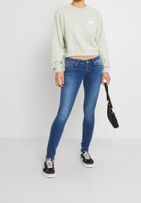 Žena v svetle zelené mikině Levi's, modrých skinny džínech, černých teniskách, držící malou černou kabelku před jednobarevným pozadím.