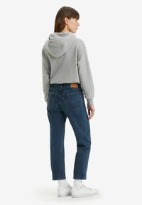 Felpa grigia con cappuccio, vestibilità comoda, abbinata a jeans blu scuro. Sneakers bianche con polsini a costine. La vista posteriore mostra i dettagli e le texture del design.