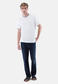 Weißes T-Shirt mit kurzen Ärmeln und schwarzem Besatz, kombiniert mit dunkelblauen Jeans und schwarzen Sandalen. Das Model steht frontal, die Hände entspannt.