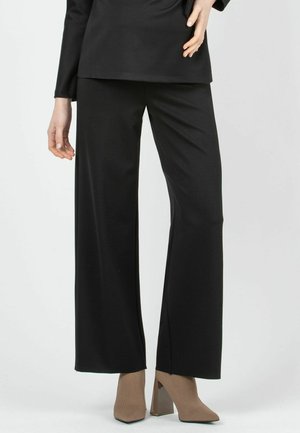 Pantalones anchos negros con textura suave, que presentan una cintura alta y líneas limpias. Combinados con zapatos de tacón cuadrado de color marrón claro.