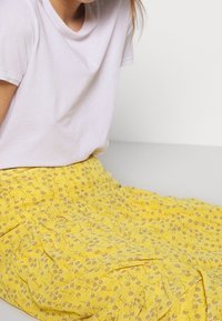 Kvinde iført en hvid kortærmet skjorte og en gul nederdel med blomsterprint, siddende mod en ensfarvet lys baggrund.