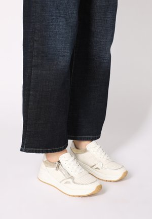 Weiße Sneakers mit Mesh- und metallischen Akzenten, ausgestattet mit einer robusten Sohle und Schnürdesign. Getragen mit dunkelblauen, weiten Jeanshosen.