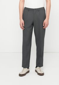 Άτομο που φοράει γκρι παντελόνια ραμμένα στα μέτρα και εκρού αθλητικά παπούτσια, στέκεται μπροστά σε λευκό φόντο.