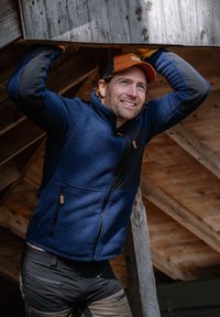 Man wearing orange cap and blue jacket lifts wooden beam inside unfinished wooden structure.