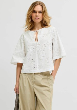 Femme aux cheveux blonds portant un chemisier blanc ajouré à manches amples et un pantalon plissé beige, tenant un sac gris dans la main droite.