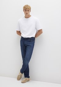 T-shirt bianco a maniche corte abbinato a jeans blu in denim e scarpe beige con lacci, in piedi davanti a uno sfondo bianco con un design minimalista.
