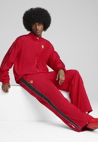 Rdeč trenirka iz lahkotnega materiala z črnimi črtami in logotipom Ferrari, ki vključuje zadrgo jakno in ohlapne hlače.