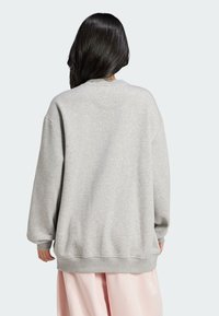 Sweatshirt gris à coupe décontractée, épaules tombantes, texture douce et ourlet côtelé. Porté sur un vêtement rose clair, mettant en avant la simplicité du design.