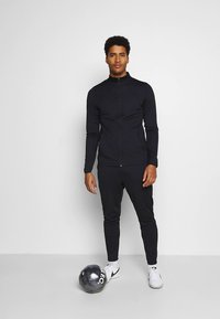 Zwarte sportieve zip-up jas met lange mouwen, gecombineerd met bijpassende broek. Draagt witte sportschoenen, staat naast een zwarte voetbal.