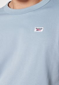 Světle modré bavlněné tričko s kulatým výstřihem, na levé horní straně má malou bílou nášivku s logem Reebok. Hladká textura.