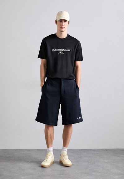 T-shirt noire à manches courtes avec logo blanc "Emporio Armani", associé à un short bleu marine et des baskets claires, avec une coupe décontractée et des poches latérales.
