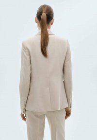 Blazer beige clair sur mesure avec une fente unique, deux coutures arrière, et des boutons décoratifs sur les manches. Tissu lisse et design structuré.