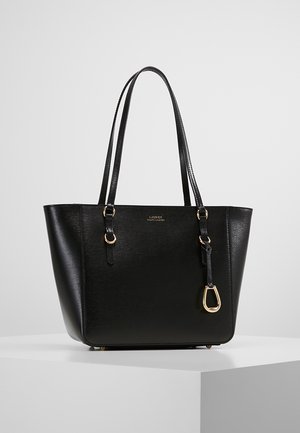 Sac fourre-tout en cuir noir avec double anse, garnitures dorées et un petit pendentif doré suspendu, présenté sur un socle blanc.