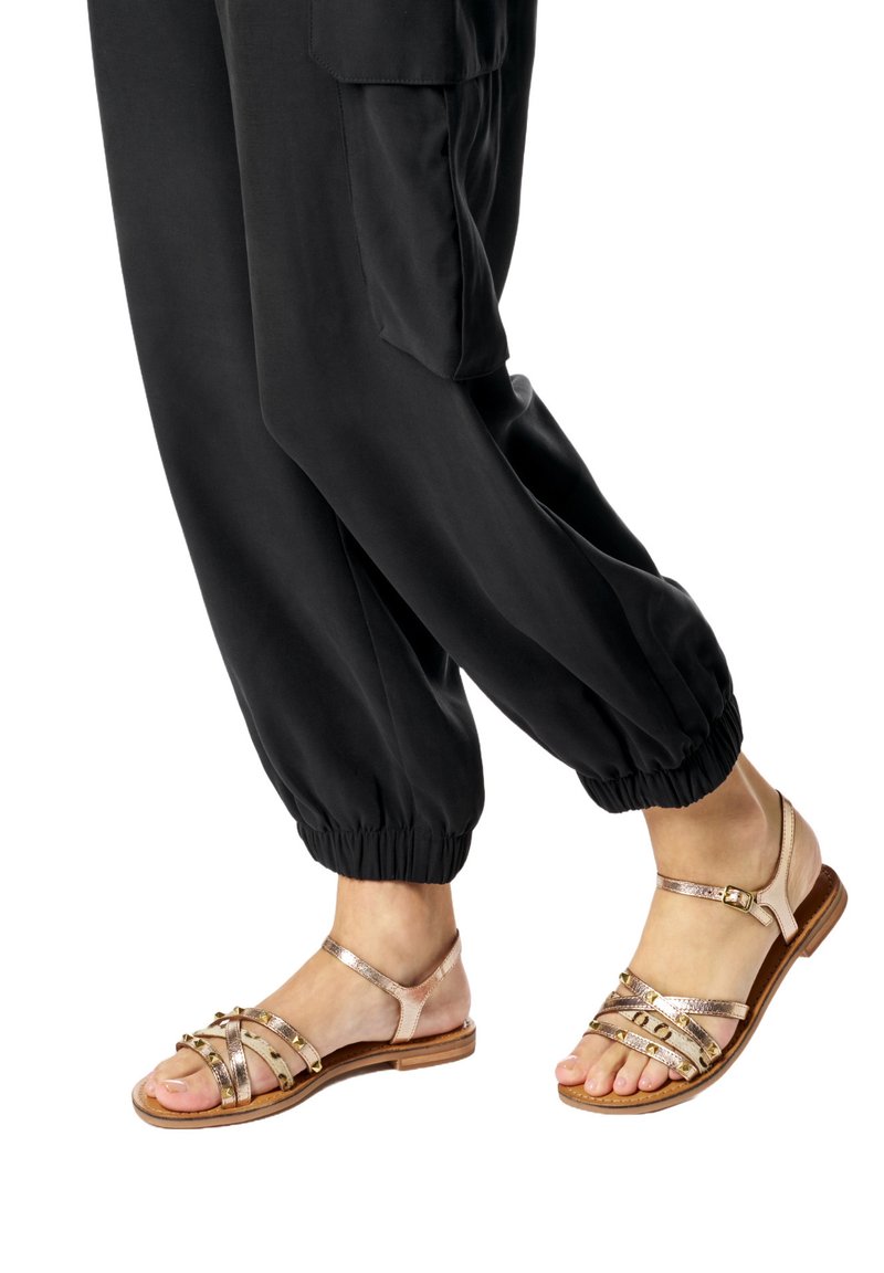 Feet wearing rose gold studded sandals with ankle straps paired with black cuffed pants.