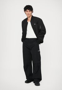 Tânăr îmbrăcat cu o jachetă bomber neagră, un tricou alb, pantaloni cargo negri largi și adidași negri, stând cu mâinile în buzunare.