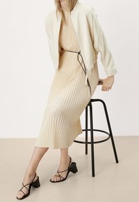 Giacca bomber crema sopra un vestito beige a pieghe, con vita strizzata e tacchi neri con cinturini, seduta su uno sgabello nero.