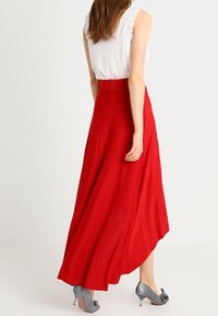 Falda maxi plisada roja con un dobladillo asimétrico. Cuenta con una cintura alta y una silueta fluida, combinada con tacones metálicos grises.