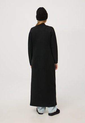 Donna che indossa un lungo cappotto nero, jeans azzurri chiari, scarpe nere e un cappello di maglia nero, in piedi di spalle contro uno sfondo uniforme.