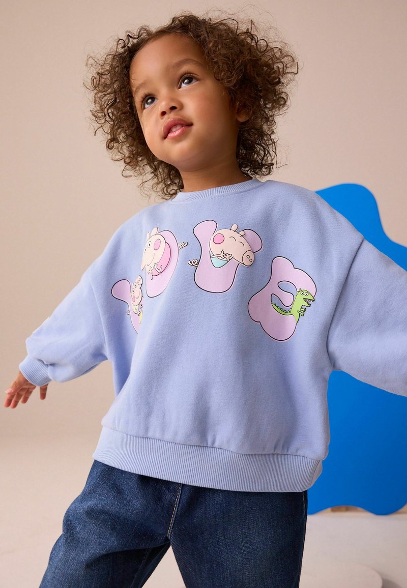 Jeune enfant aux cheveux bouclés portant un sweat-shirt bleu clair avec des personnages de dessins animés et un jean bleu, posant avec les bras légèrement écartés.