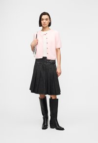 Γυναίκα που φοράει ανοιχτό ροζ κοντομάνικο καρντιγκάν πάνω από λευκή μπλούζα, μαύρη πλισέ φούστα με ζώνη, μαύρες μπότες μέχρι το γόνατο και κρατάει μια ασημένια τσάντα.