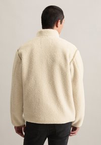 Crème-kleurige fleece jas met een hoge kraag, gestructureerd oppervlak en lange mouwen, gezien van de achterkant. Zwarte jeans maken de look compleet.