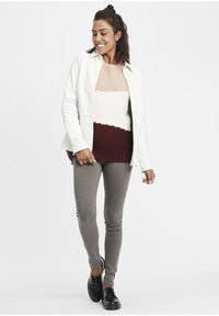 Pull en tricot superposé avec des rayures beige, crème et bordeaux ; associé à un jean skinny gris et des chaussures noires. Veste extérieure blanche.