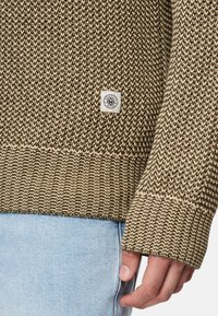 Stickad tröja i olivgrönt och beige, med ett texturerat mönster, ribbat avslut och en fyrkantig logotyp på sidan.