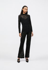 Salopetă din dantelă neagră cu mâneci lungi, model floral, corset ajustat și pantaloni evazați cu fante laterale. Tocuri negre cu ținte.