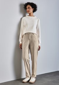Beige texturierte Hose, weißes Langarmoberteil mit U-Boot-Ausschnitt, kombiniert mit weißen und beigen Sneakers, stehend vor einer neutralen Wand.