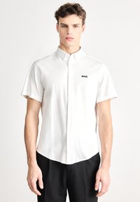 Baltas trumpomis rankovėmis švarkas su sagomis ir išlenktu apdailos kraštu, kairėje krūtinės pusėje mažas juodas logotipas „BOSS“. Lygus audinio paviršius.