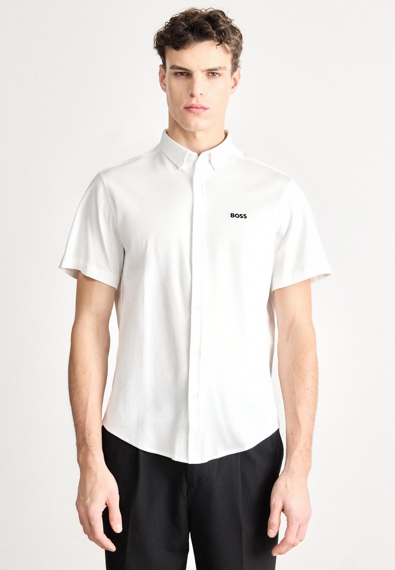 Baltas trumpomis rankovėmis švarkas su sagomis ir išlenktu apdailos kraštu, kairėje krūtinės pusėje mažas juodas logotipas „BOSS“. Lygus audinio paviršius.