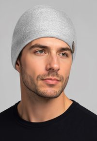 Grijze gebreide muts met een gladde textuur en een klein bruin logolabel, gedragen door een persoon in een zwart shirt. Close-up van het gezicht.