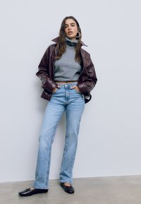 Žena v hnedej koženej bunde, šedej roláku, svetlotyrkých džínsoch a čiernych baletných topánkach, stojí oproti jednoduchému múru.