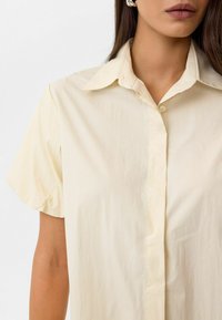 Lichtbeige kortemouwen shirt met een klassieke kraag, knopen voorop en een gladde, licht gerimpelde structuur. Eenvoudig en minimalistisch ontwerp.