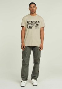 Beige katoenen t-shirt met vet zwart tekst, gecombineerd met groene cargobroek met meerdere zakken en een gestructureerde stof. Witte sneakers maken de look compleet.