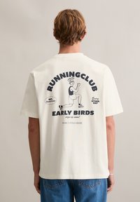 Licht crèmekleurig katoenen T-shirt met een grafische print op de achterkant met tekst en een cartoonfiguur, gecombineerd met blauwe spijkerbroek.