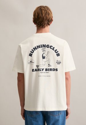 Jasnokremowy bawełniany t-shirt z graficznym nadrukiem na plecach, zawierającym tekst i postać z kreskówki, zestawiony z niebieskimi jeansami.