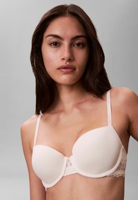 Joven mujer con cabello largo y oscuro que lleva un sujetador acolchado beige claro con detalles de encaje sobre un fondo gris liso.