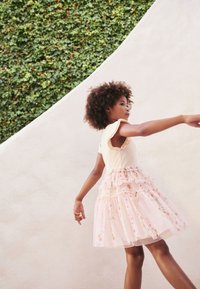 Licht beige top met geruite mouwen gaat over in een gelaagde, roze tule rok versierd met bloemenpatronen en doorschijnende accenten.