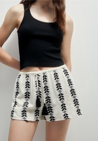 Femme portant un haut court noir sans manches et un short blanc avec des motifs géométriques noirs verticaux et des cordons à pompons.