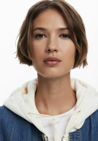 Model wears a denim jacket with a white hooded sweatshirt. The short hair frames the face, featuring neutral skin tones and minimal makeup.