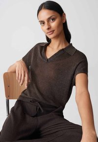 Femme aux longs cheveux foncés portant un haut en maille marron à manches courtes avec col et un pantalon assorti, assise sur une chaise en bois devant un fond uni.