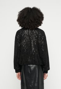 Juodas sekančių bombonešio stiliaus švarkelis su tekstūruotu paviršiumi, ribuotais rankogaliais ir laisvu kirpimu; matomas iš nugaros kartu su juoda odine suknele.